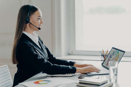 Attractive businesswoman in formal suit wearing headset having video call on laptop computer, working with graphs charts, side view. Satisfied young female economist presenting financial report onlineの写真素材
