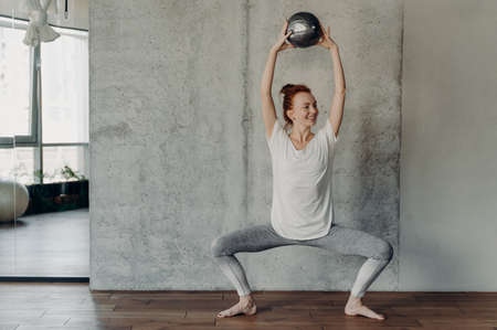 Full leghth shot of slim happy ginger female performing barre exercises with small fitball lifted up...