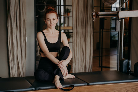 Beautiful ginger woman in sportswear sitting on Cadillac machine with arms around legs in calm and relaxed position after exercising in gym or fitness studio. Sport and healthy lifestyleの写真素材