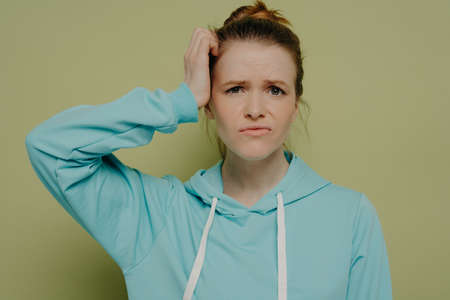 Young puzzled female student in blue hoodie expressing confused emotion, holding hand on head and...