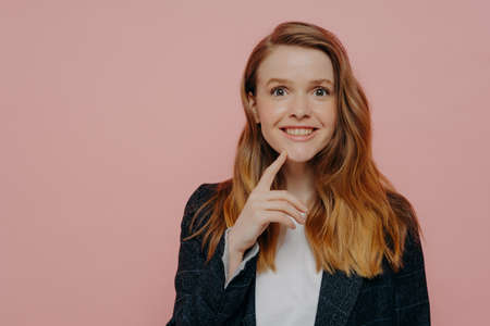 Young excited ginger lady in jacket with white shirt, realizing there is solution, generating new idea with finger on chin, standing alone next to pink background and expressing happinessの写真素材