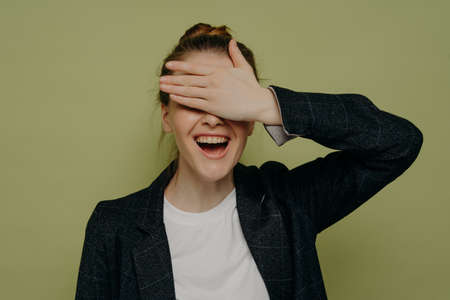 Laughing young female in casual clothes covering eyes with hand while waiting for surprise, smiling teenage girl keeping mouth opened with embarrassed expression, studio shotの写真素材