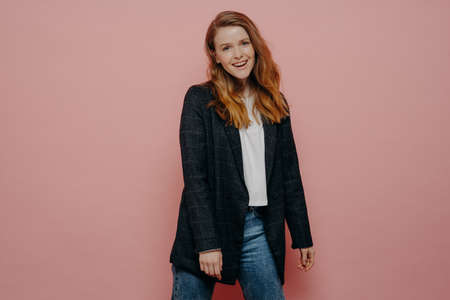 Stylish fashionable young woman wearing plaid blazer and jeans posing in studio, feeling carefree and expressing positive emotions while posing isolated over pink wall background. Happy people conceptの写真素材