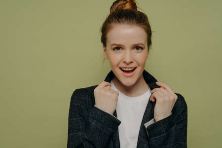 Headshot portrait of young overjoyed trendy woman in casual clothes holding collar of jacket and looking at camera with excited face expression, positive female posing in studio over green backgroundの写真素材