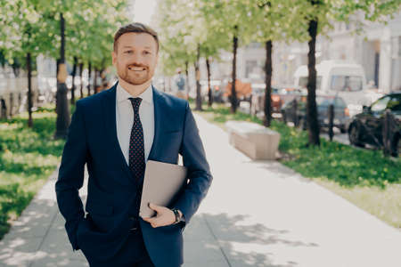 Relaxed happy handsome male office worker in blue suit holding laptop with important data and presentation, taking walk in park while taking break from work, enjoying fresh air and greeneryの写真素材