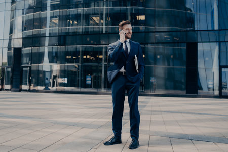Handsome young bearded office worker in dark blue formal suit speaks on cellphone while standing outside next to business center waiting for partners to meet. Business people conceptの写真素材