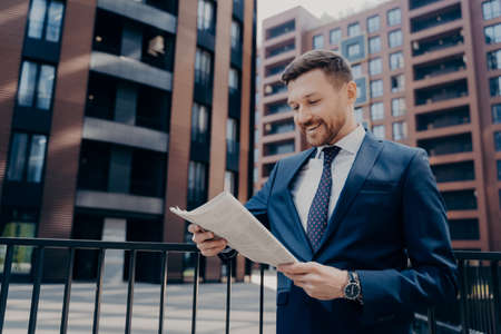 Successful young male economist reading business press in urban setting background, being happy with financial news in morning newspaper while holding it in hands. Bussinessman in formal suit outdoorsの写真素材