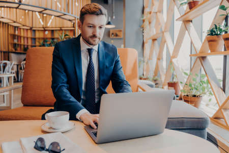 Young confident businessman in formal suit works online on laptop in lobby of corporate center during coffee break, preparing presentation for business meeting with new partnersの写真素材