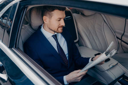 Thoughtful young bearded goodlooking executive manager in formal dark blue suit reading newspaper in...
