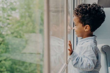 Side view of lovely afro american kid toddler waiting for mom, dressed in blue shirt, looking out of window through his reflection leaning on glass with hands and smiling. Children leisure time indoorの写真素材