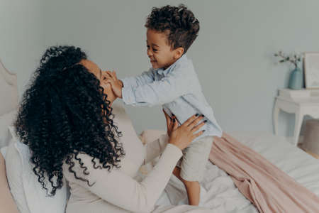 Happy afro american family, pregnant woman loving mom in white dress with curly hair sitting on white bed and joyfully spending time with child, giving attention to her son and playing with himの写真素材