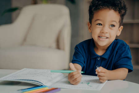 Cute mixed race small boy in polo tshirt painting coloring book with felt tip pens, sitting by table...