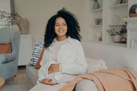 Can not wait to become mom. Positive smiling pregnant afro american woman in knitted beige dress touching gently her belly and looking with smile aside at home. Pregnancy and expectation conceptの写真素材