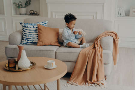 Photo of living room with small lovely mulatto child peacefully sitting on sofa among pillows and coverlid in foreground of coffee table with teacup and vases decorations, kid playing at homeの写真素材