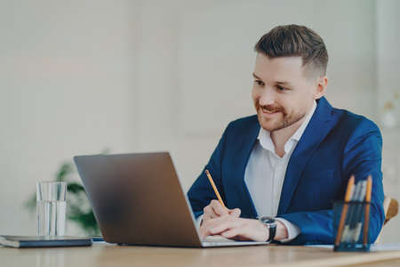 Corporate worker in formal suit sits at desktop focused at laptop computer poses in office interior...