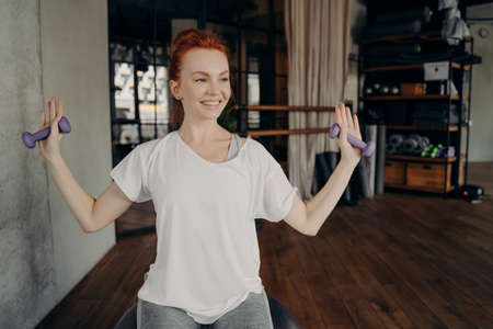 Happy sporty european redhead young woman sits on silver fitball and raises arms with small dumbbells during workout in fitness studio. positive female in active wear with sport equipment in gymの写真素材