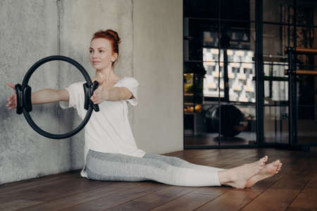 Young active red haired lady sitting with legs stretched forward and holding pilates ring by both hands performing strength exercises in fitness studio or gym. Concept of healthy lifestyle and sportの写真素材