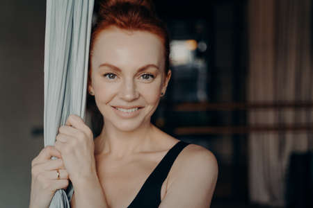 Close up portrait of young beautiful fit lady with red hair holding hammock, enjoying antigravity...