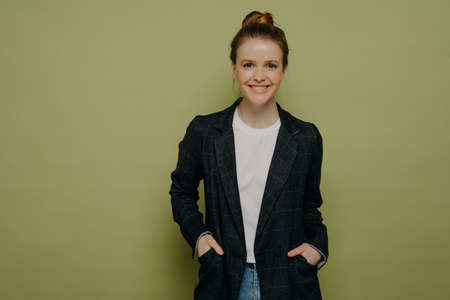 Smiling confident teenage girl with hair in bun wearing stylish plaid blazer and jeans keeping hands in pockets and looking at camera with happy expression while standing against green wallの写真素材