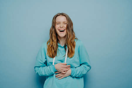 Overjoyed young woman dressed in hoodie holding belly and cant stop laughing after hearing hilarious joke, keeping eyes closed and having fun while standing isolated over blue background, studio shotの写真素材