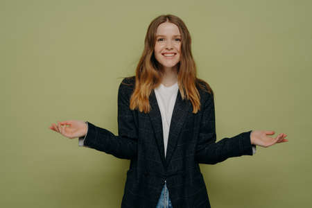 Smiling positive female shrugging shoulders with spreaded hands and looking at camera with positive...