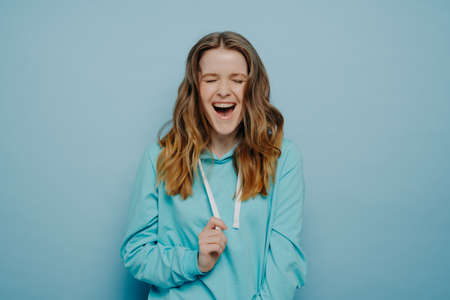 Portrait of optimistic teenage girl in casual clothes with tongue piercing keeping eyes closed and laughing joyfully, expressing happiness while posing isolated over blue background in studioの写真素材
