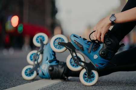 Cropped shot of unrecognizable woman puts on rollerblades poses outdoors on asphalt against blurred...
