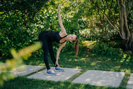 Active fitness model exercises outdoors leans to feet raises arm prepares muscles before cardio training challenges herself dressed in active wear has pony tail poses in green park during sunny dayの写真素材