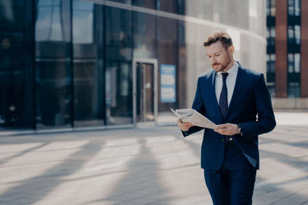 Focused bearded young businessman in formal outfit walking alone with newspaper in hands, reading latest news on his way to workplace with office buildings in backgroundの写真素材