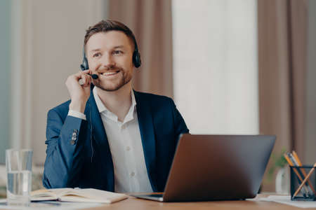 Photo of happy male manager wears headset works on laptop computer consults clients looks with glad...