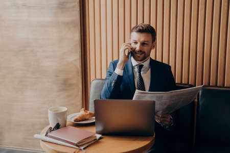 Satisfied business owner in blue suit talking on phone with newspaper in hand, sharing recent news with his partner and looking at laptop, notifying about further developments while working in cafeの写真素材