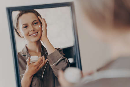 Positive happy young woman in pajama smiling, applying facial cream and looking in mirror, attractive lady doing morning beauty routine in bedroom at home. Skin care conceptの写真素材