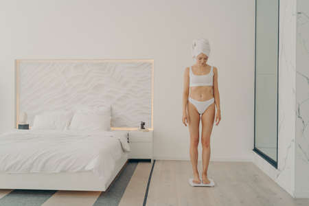 Beautiful young good looking female in underwear with head wrapped in towel looking down while standing in bedroom on scale and measuring her weight. Beauty and body care conceptの写真素材