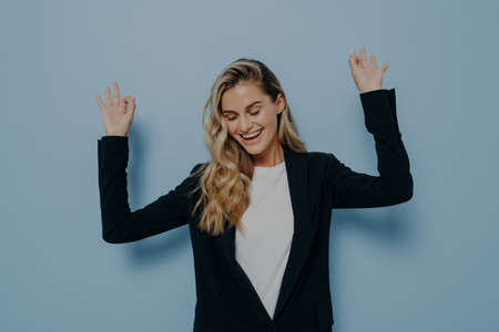 Portrait of carefree happy woman with blonde dyed hair raising her arms and dancing with closed eyes and smile on face while standing isolated against blue background. Happiness conceptの写真素材