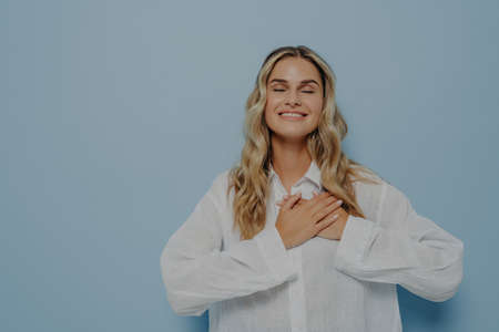Pleased happy thankful woman with blond wavy hair in white oversize shirt gesturing appreciation and...