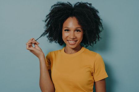 Studio portrait of beautiful young dark skinned woman with shaggy hairstyle smiling cheerfully, showing white teeth at camera, while playing with lock of her hair, feeling happy and excitedの写真素材