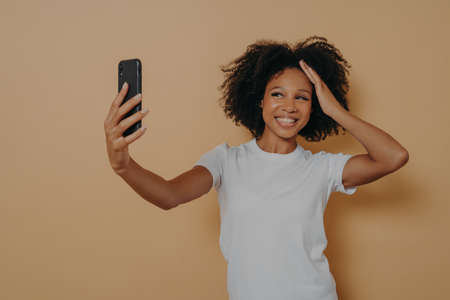 Happy charming dark skinned female 20s in white tshirt making photo selfie on modern smartphone and smiling cheerfuly, isolated over beige wall. Positive young african woman taking photo on cellphoneの写真素材