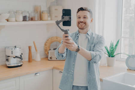 Young man blogger recording video blog or live streaming with smartphone and gimbal indoors standing in kitchen background. Social media vlogger shooting vlog, streaming online podcast on mobile phoneの写真素材