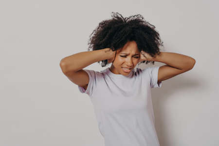 Studio portrait of young angry annoyed african woman covering her ears with both hands to avoid loud...