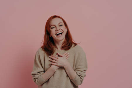 Ha-ha. Delighted red-haired funny woman laughing out loud, dressed in beige sweater, holding both hands on chest and smiling cheerfully, posing against pink background with copy space for textの写真素材