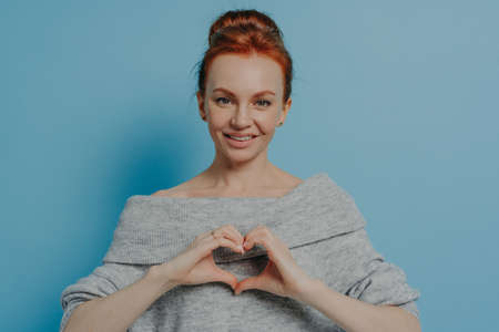Kind smiling red haired woman showing heart gesture on chest while standing isolated on blue wall background, happy ginger female demonstrating love, care and tenderness, dressed in knitted sweaterの写真素材