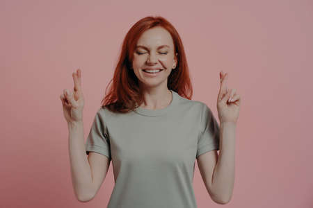 Studio shot of hopeful red-haired female student with closed eyes in casual wear crossing fingers...