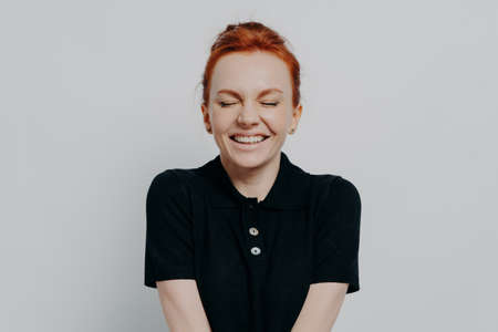 Beautiful and happy pleased red haired woman with closed eyes and toothy smile posing against grey wall. Cheerful excited female demonstrating positive emotions and happiness, dressed casuallyの写真素材