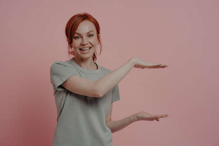 Young pleasant red-haired woman in t-shirt showing huge product size or something big with hands and smiling while standing isolated on pink background. Promotion and advertising conceptの写真素材