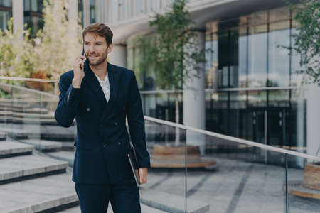 Caucasian satisfied man in formal outfit sitting on steps outdoor, talking on mobile phone and...