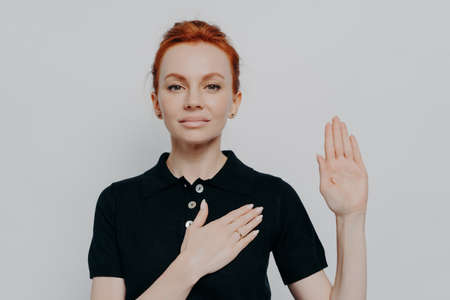 Beautiful red-haired patriotic woman wearing black t-shirt swearing with hand on chest and open palm, making loyalty promise oath while posing isolated over grey studio backgroundの写真素材