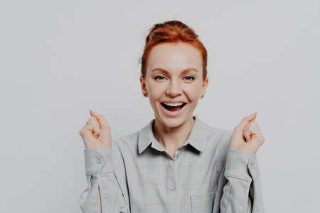 Happy red haired 30s woman wearing casual shirt holding clenched fists and looking at camera with...