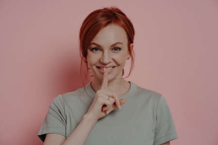Young beautiful smiling red-haired woman telling secret information, posing isolated over pink wall,...