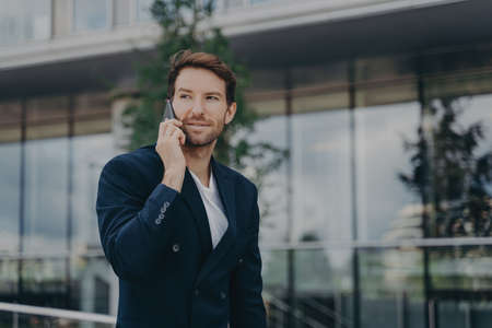 Male entrepreneur talks via phone solves urgent problems poses near business center dressed in black suit uses modern technologies for communication looks away thoughtfully. Technology work lifestyleの写真素材