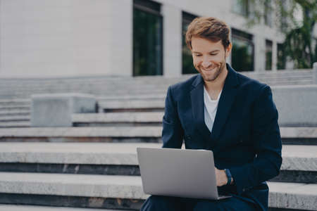 Young businessman sits on steps uses laptop organizes meeting online with investors has happy expression uses free internet poses outdoor dressed formally. Corporate director analyzes web informationの写真素材